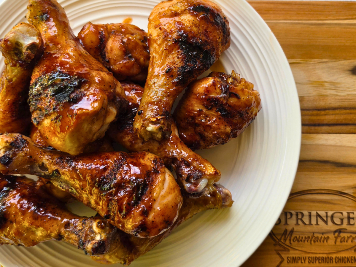 Smoky BBQ grilled chicken drumsticks piled on a plate, coated in a glossy barbecue sauce with visible char from the grill, served on a wooden board branded with Springer Mountain Farms