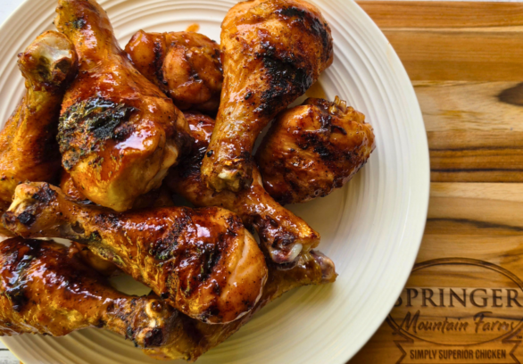 Smoky BBQ grilled chicken drumsticks piled on a plate, coated in a glossy barbecue sauce with visible char from the grill, served on a wooden board branded with Springer Mountain Farms