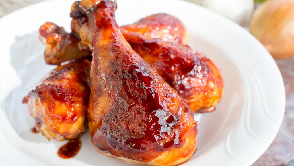 Grilled BBQ chicken drumsticks glazed with sauce on a white plate, with fresh garlic and onion in the background