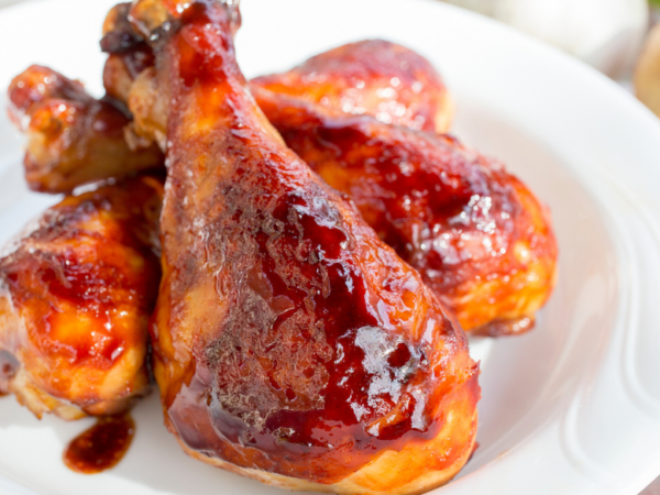 Grilled BBQ chicken drumsticks glazed with sauce on a white plate, with fresh garlic and onion in the background