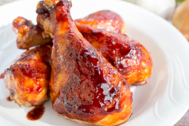 Grilled BBQ chicken drumsticks glazed with sauce on a white plate, with fresh garlic and onion in the background